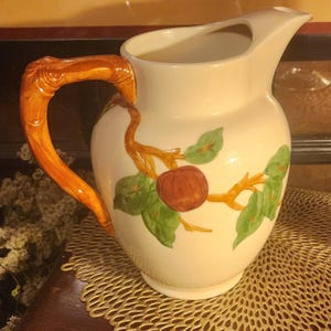 May include: Cream-colored ceramic pitcher with a decorative orange handle. The pitcher features a painted design of a red apple, green leaves, and orange branches. A classic piece for serving beverages.