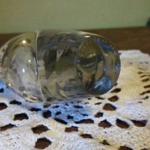 May include: A clear glass bottle stopper with a textured surface and a circular design on the bottom. The design is in a dark gray color and appears to be a logo or inscription.
