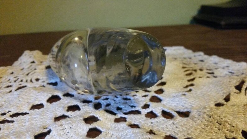 May include: A clear glass bottle stopper with a textured surface and a circular design on the bottom. The design is in a dark gray color and appears to be a logo or inscription.