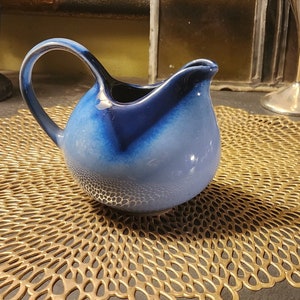 May include: A blue and white ceramic creamer with a rounded shape and a handle. The creamer is sitting on a gold-colored placemat.