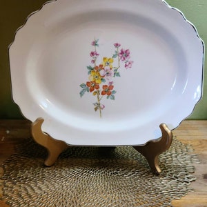 May include: A white oval serving platter with a scalloped edge and a floral design in shades of pink, yellow, and orange. The platter is on a gold metal stand.