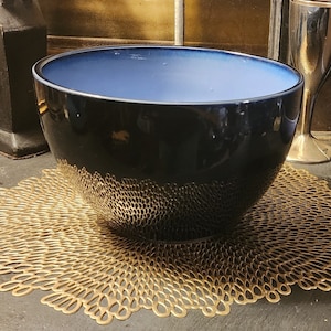 May include: A large, dark blue ceramic bowl with a light blue interior. The bowl is sitting on a gold-colored, decorative placemat.