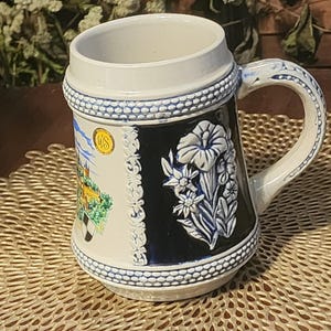 May include: A ceramic beer stein with a white and blue design. The stein features a handle and a detailed relief of flowers and a castle scene. The stein has a white rim and base with blue accents. A gold emblem is visible on the side.