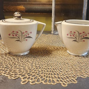 May include: A white sugar bowl and creamer set with a floral design. The set features a delicate floral pattern with pink flowers and gray leaves. The sugar bowl has a lid and both pieces have a silver trim.