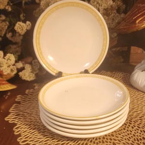 May include: A stack of white ceramic plates with a yellow Greek key border. One plate is standing upright behind the stack. The plates are on a gold-colored, decorative placemat. The background is dark and out of focus.