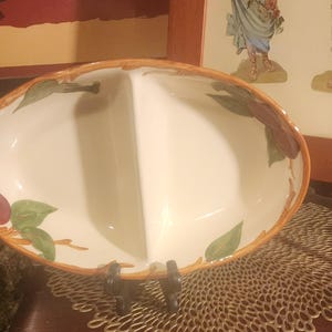 May include: Oval, two-section serving dish with a white interior and a decorative rim. The rim is brown with painted green leaves and red fruit accents. The dish is supported by a black metal stand.