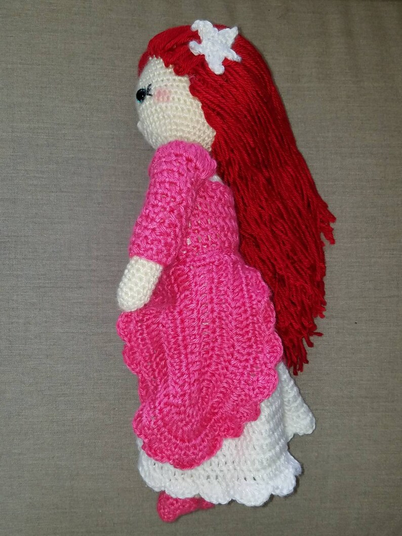 May include: A crocheted doll with long red hair, a pink dress, and a white flower in her hair.