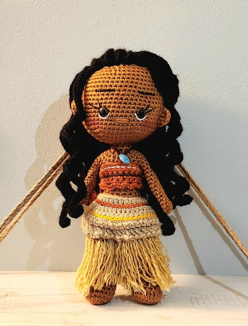 May include: Handmade crochet doll with a brown complexion, black curly hair, and a detailed outfit. The doll is dressed in a brown and orange top, a tan skirt with yellow details, and a fringed bottom. A blue pendant is visible.