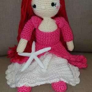 May include: A crocheted doll with red hair, a pink dress, and a white crocheted skirt. The doll is holding a white starfish.