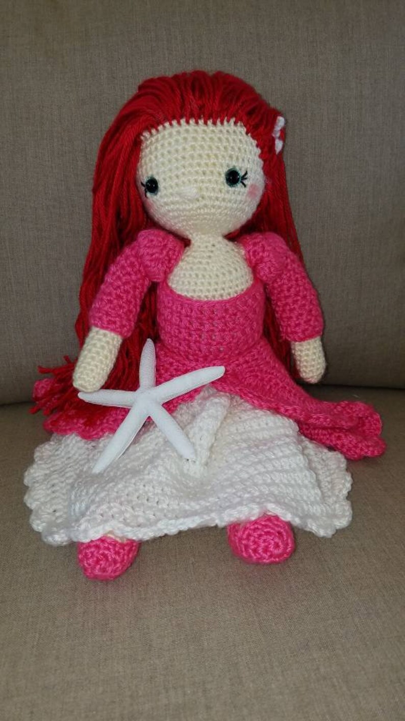 May include: A crocheted doll with red hair, a pink dress, and a white crocheted skirt. The doll is holding a white starfish.