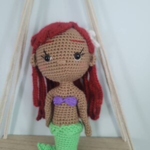 Puede incluir: Mu&ntilde;eca de sirena de crochet con cabello rojo, piel marr&oacute;n, top morado y cola verde.