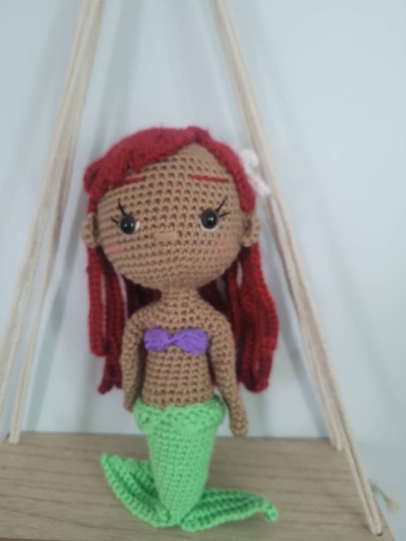 Puede incluir: Mu&ntilde;eca de sirena de crochet con cabello rojo, piel marr&oacute;n, top morado y cola verde.