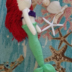 May include: A crocheted mermaid doll with red hair, a white bodice, a purple seashell bra, and a green tail. The doll is lying on a blue and brown rug with a starfish and seashells.