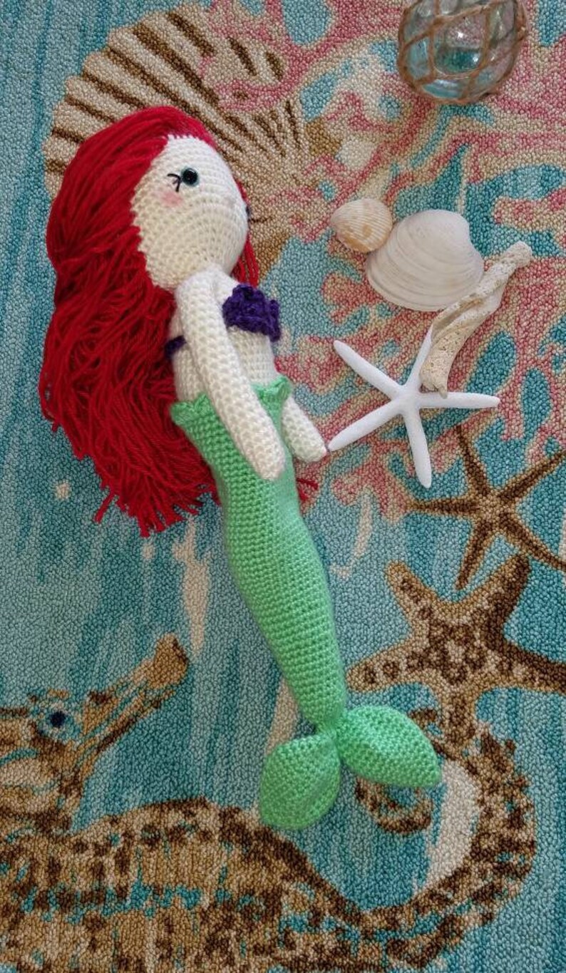 May include: A crocheted mermaid doll with red hair, a white bodice, a purple seashell bra, and a green tail. The doll is lying on a blue and brown rug with a starfish and seashells.