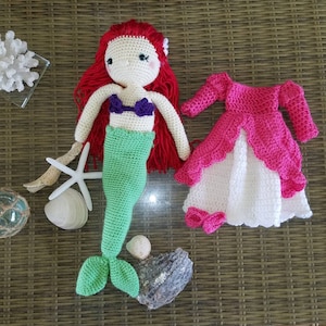 May include: A crocheted mermaid doll with red hair, a green tail, and a purple seashell bra. The doll is wearing a pink and white crocheted dress.
