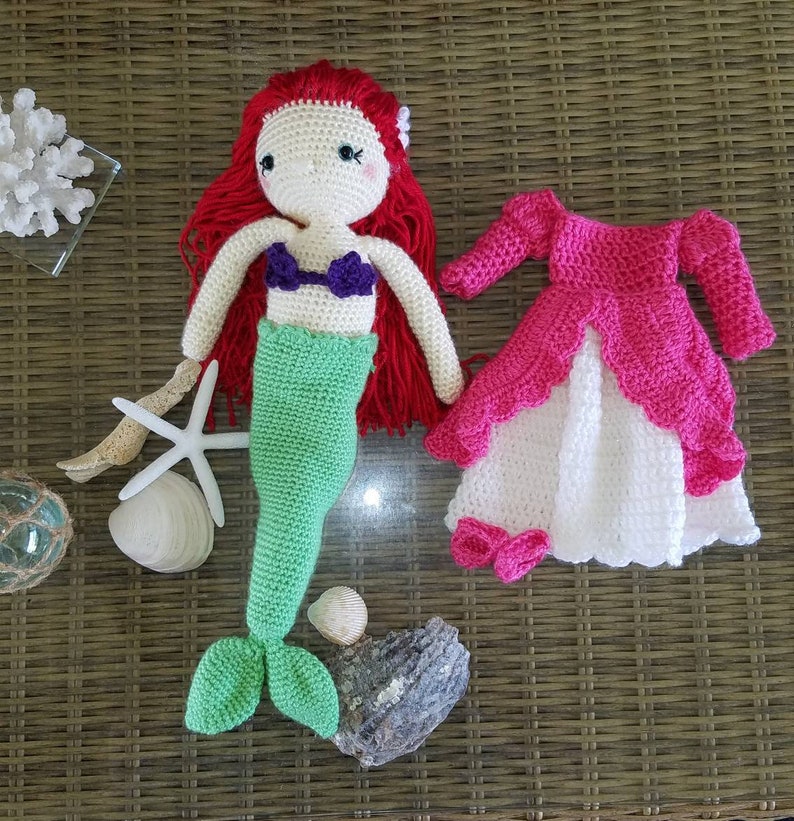 May include: A crocheted mermaid doll with red hair, a green tail, and a purple seashell bra. The doll is wearing a pink and white crocheted dress.
