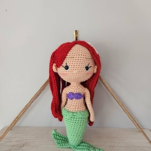 Puede incluir: Una mu&ntilde;eca de sirena de crochet con cabello rojo, un sujetador de concha p&uacute;rpura y una cola verde.