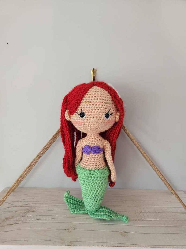 Puede incluir: Una mu&ntilde;eca de sirena de crochet con cabello rojo, un sujetador de concha p&uacute;rpura y una cola verde.