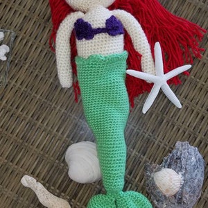 May include: Crocheted mermaid doll with red hair, a purple seashell top, and a green tail. The doll is holding a white starfish.