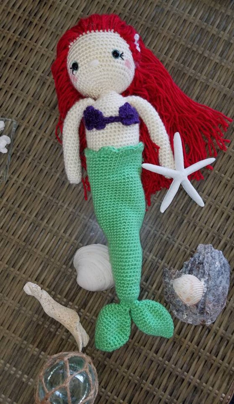 May include: Crocheted mermaid doll with red hair, a purple seashell top, and a green tail. The doll is holding a white starfish.