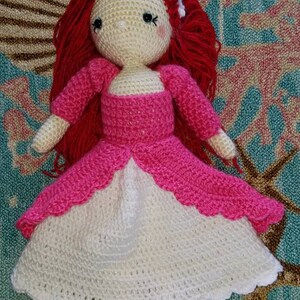 May include: A crocheted doll with red hair wearing a pink and white dress. The doll is sitting on a colorful rug.