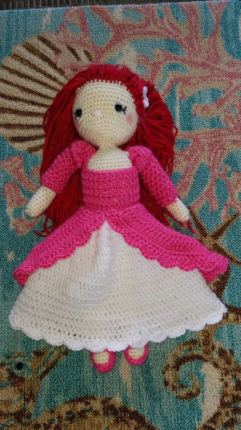 May include: A crocheted doll with red hair wearing a pink and white dress. The doll is sitting on a colorful rug.