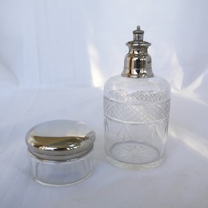 May include: Clear glass perfume bottle with a silver-colored stopper and a matching small jar with a silver lid. The perfume bottle has a decorative diamond pattern around the middle. The items are on a white surface.