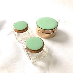 Könnte beinhalten: Drei Vintage-Kosmetikdosen mit mintgrünen Deckeln und goldfarbenen Rändern. Zwei Dosen sind quadratisch, eine rund. Die Dosen bestehen aus klarem Glas, die Deckel sind pastellgrün.