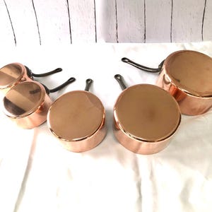 May include: A collection of copper pots with black handles. The pots vary in size and are arranged on a white surface. The copper has a shiny, reflective surface, and the handles are curved.
