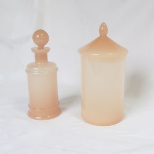 May include: Two peach-colored glass containers. One is a tall, cylindrical jar with a conical lid. The other is a bottle with a rounded base and a stopper. Both have a frosted appearance and are set against a white background.