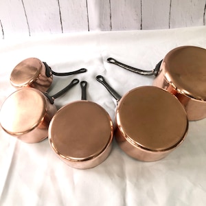 May include: A collection of copper pots and pans with black handles. The cookware is arranged on a white surface, showcasing their shiny, reflective surfaces. The set includes various sizes, suitable for different cooking needs.