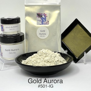 May include: A white powder pigment called Gold Aurora #501-IG in a black bowl. The pigment is also shown in a small jar and a larger bag. The product is used for creating a pearlescent effect in paint, resin, and other mediums.