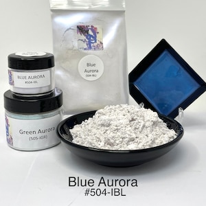 May include: Two small jars of iridescent pigment powder, one labeled "Blue Aurora #504-1BL" and the other "Green Aurora (505-IGR)". A larger bag of white pigment powder labeled "Blue Aurora (504-1BL)" is in the background. A small square dish of blue pigment powder is also in the background.