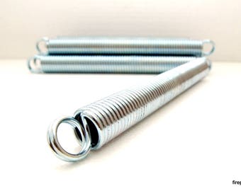 May include: Close-up of several silver metal extension springs. One spring is in focus, with a loop at each end. Two other springs are slightly out of focus in the background. The springs are on a white surface.