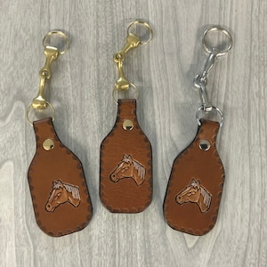May include: Three brown leather keychains with a horse head design. Each keychain has a different metal ring attachment. The keychains are on a woodgrain surface.