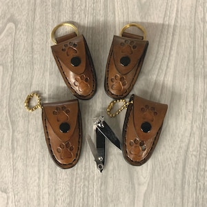 May include: Four brown leather keychains with paw print designs and a pair of nail clippers. The keychains have a snap closure and a gold-colored key ring or chain. The nail clippers are silver and black.