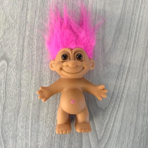 May include: A vintage troll doll with a smiling face, black eyes, and bright pink hair. The doll has outstretched arms and a small pink belly button. The doll is tan-colored and stands on a gray wooden surface.