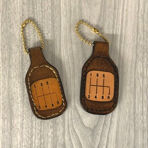 May include: Two brown leather keychains with a gold chain and a gear shift pattern. The keychains have a light brown center with the numbers 1, 2, 3, 4, 5, 6, and R. The keychains are stitched with a brown thread.