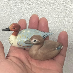 May include: Two small, hand-painted duck figurines. One duck has a brown head, white neck, and a light blue body with yellow accents. The other duck is brown with a darker brown head. Both have black beaks and are resting in a hand.