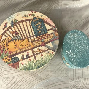 May include: Two round, decorative boxes. The larger box is cream colored with a whimsical illustration of a birdhouse, birds, and a cat napping on a bench. The smaller box is teal blue with a speckled texture.