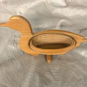 May include: A light brown wooden duck-shaped basket with a collapsible, tiered oval structure. The object features a smooth, natural wood grain finish and is designed with a flat base for stability, showcasing a carved silhouette of a duck.