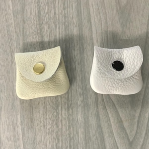 May include: Two small square pouches made of textured leather. The left pouch is cream-colored with a gold-tone metal snap button closure. The right pouch is white with a black snap button closure. Both pouches feature a rounded flap design.