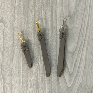May include: Three gray leather keychains with metal clasps. The clasps are gold and silver. The leather is a muted gray color. The keychains are of varying lengths, with the longest measuring approximately 6 inches.