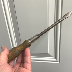 May include: A vintage flathead screwdriver with a wooden handle and a metal shaft. The handle is a light brown color, and the metal shaft shows signs of wear. The screwdriver is held in a hand.