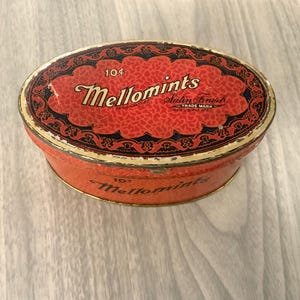 May include: Vintage oval Mellomints tin with a red and black design. The lid features the text "Mellomints" in a script font, with "10¢" above. The tin has a satin finish and a trade mark. The tin is on a light wood surface.
