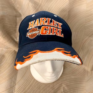 May include: A navy blue baseball cap with the words "HARLEY GIRL" in orange block letters. The brim is white with orange flame accents. The Harley-Davidson logo is also visible on the cap.