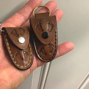 May include: Two brown leather keychains with baby footprint designs. One keychain has a silver chain, and the other has a gold ring. Both have a snap closure and stitched edges.