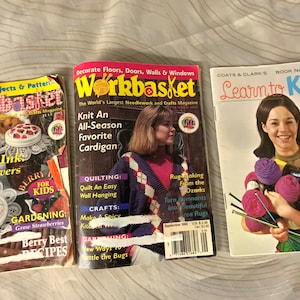 May include: Three craft magazines with colorful covers. The first magazine is titled "Workbasket" and features a summer issue with a berry theme. The second magazine is also titled "Workbasket" and features a knit cardigan. The third magazine is titled "Learn to Knit" and features a woman holding balls of yarn.