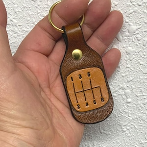 May include: A brown leather keychain featuring a gear shift pattern with numbers 1 through 6 and reverse (R). The keychain has a gold-colored ring and a brass stud. The leather is a medium brown color.