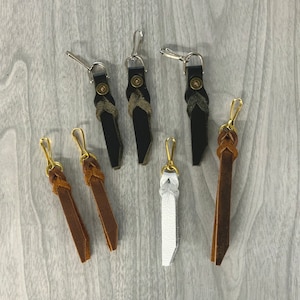 May include: A collection of leather keychains in various colors, including black, brown, and white. Each keychain features a metal clasp and decorative braided or stitched leather details. The keychains are arranged on a light gray wooden surface.